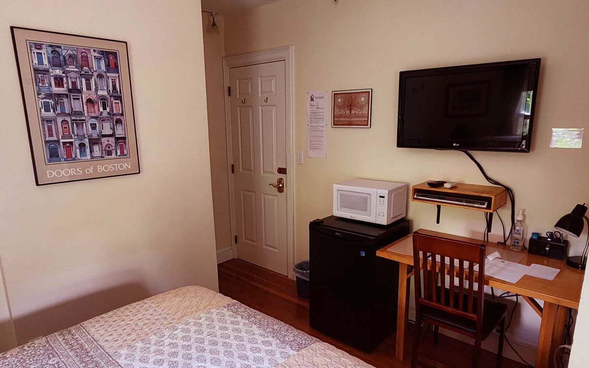 corner of queen bed with quilt, TV, table, fridge,microwave.
